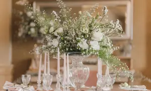Eleganckie ustawienie stołów na 20 osób. Centralny punkt stanowi bukiet białych kwiatów w szklanym wazonie, otoczony świecami i kryształowymi kieliszkami.