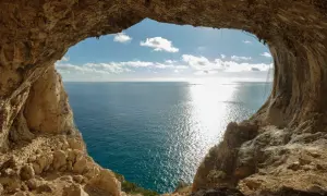 Vista dal mare da una grotta, con un sentiero del pellegrino che si intravede tra le rocce. Il sole brilla sull'acqua.