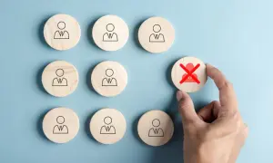 Dłoń wybiera drewniany krążek z przekreśloną postacią, symbolizującą rejestrację jako bezrobotny.