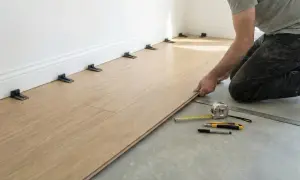 A person kneels, laying a light wood vinyl plank. Spacers are placed along the wall, showing the best way to lay vinyl plank flooring. Tools like a tape measure and ruler are nearby.