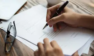Dłoń pisze na dokumencie, który może być związany z art. 30 § 1 pkt 1 kodeksu pracy. Obok leżą okulary i tablet.