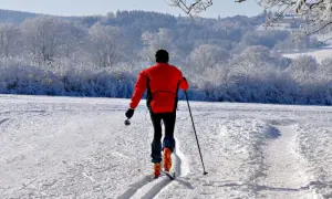 Narciarz w czerwonej kurtce przemierza przygotowane trasy biegowe narciarskie. W tle mroźny, zimowy krajobraz z ośnieżonymi drzewami.