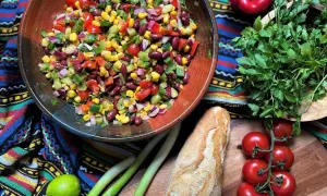Bunter mexikanischer Salat mit Mais, Bohnen und Gemüse in einer Schüssel, daneben Baguette, Limette und Kräuter.