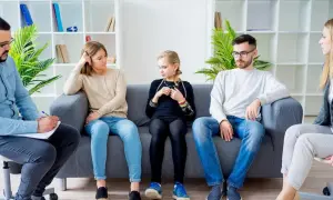 Rodzina podczas terapii systemowej. Terapeuta notuje, rodzice i córka siedzą na kanapie, każdy pogrążony we własnych myślach.