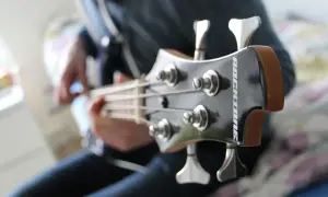 Osoba gra na gitarze basowej. Widoczna jest główka instrumentu z kluczami i strunami.