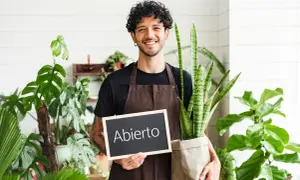 Joven sonriente con plantas, sosteniendo un cartel de "Abierto". Inspiración para ideas de negocios para jóvenes en el sector verde.