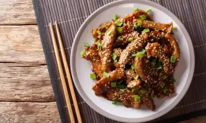 Pyszny kurczak teriyaki z sezamem i szczypiorkiem na talerzu, obok pałeczki.