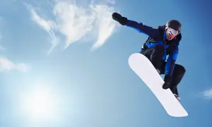 Snowboardzistka w locie na tle błękitnego nieba. Jej deska snowboardowa damska jest biała i lśniąca.