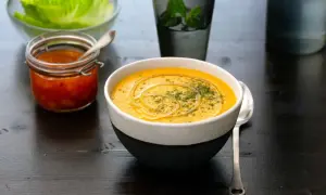 En krämig linssoppa med en virvel av olja och örter. Bredvid står en burk med en röd röra och en skål med salladsblad. Ett perfekt linssoppa recept för en mysig kväll.
