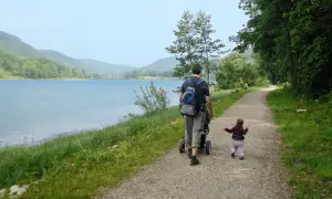 Vater schiebt Kinderwagen am Happurger Stausee entlang, während das Kind vorausläuft. Herrliche Kulisse für eine Wanderung.