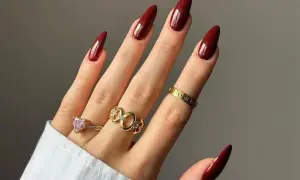 Main d'une femme aux ongles longs et pointus, vernis d'un bordeaux profond, parfaite tendance ongle automne. Bagues dorées et une bague cœur rose ornent ses doigts.
