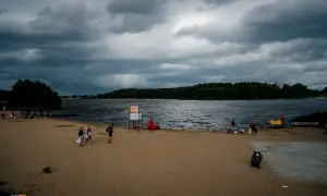 Plaża nad jeziorem Wysoka, z ciemnymi chmurami na niebie. Ludzie spacerują po piasku, niektórzy odpoczywają. W tle las i spokojna tafla wody.
