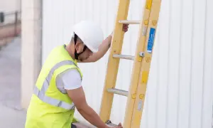 Pracownik w kamizelce odblaskowej i kasku przygotowuje drabinę. Czy można wykonywać pracę na drabinie bez potrzebnych kursów wysokościowych? Bezpieczeństwo przede wszystkim!