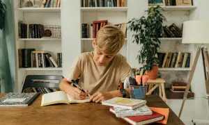 Un niño concentrado escribe en su cuaderno, rodeado de libros y útiles escolares, practicando dictados cortos para mejorar su ortografía.