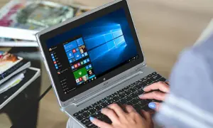 Kobieta pisze na laptopie z systemem Windows 10. W tle widać stosy czasopism, co sugeruje, że zadania w tle ochrony systemu są wykonywane podczas relaksu.