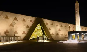 Nowe muzeum w Egipcie nocą. Monumentalna fasada z geometrycznymi wzorami i podświetlonym wejściem. Obok stoi starożytny obelisk.