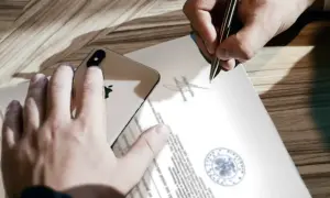 Dłoń trzyma iPhone'a na dokumencie, podczas gdy druga ręka podpisuje go, co może symbolizować poddanie się egzekucji.
