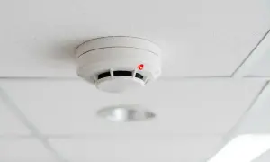 Détecteur de fumée blanc sur un plafond blanc, avec un voyant rouge allumé, suggérant un détecteur qui se déclenche tout seul.