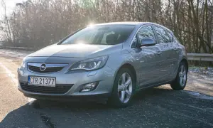 Szary Opel Astra J 1.6 benzyna na tle drzew.