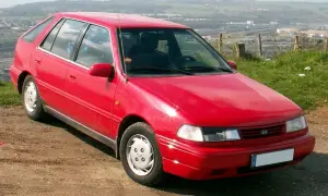 Czerwony Hyundai Pony z lat 90. stoi na brudnej drodze, z widokiem na dolinę i miasto w tle.