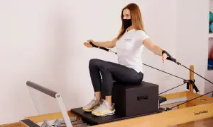 Kobieta w masce ćwiczy na reformerze pilates, wykonując ćwiczenie z linkami.