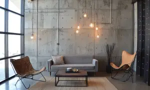 Salon w industrialnym stylu: szara kanapa, dwa fotele (jeden skórzany, drugi z brązowej skóry), drewniany stolik kawowy, betonowa ściana z widocznymi instalacjami i lampami.