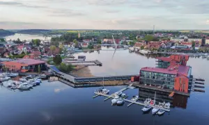 Widok na nowoczesny hotel i przystań jachtową na Mazurach. Widać liczne łodzie i żaglówki, a w tle miasteczko z charakterystyczną architekturą. To idealne mazury atrakcje dla miłośników żeglarstwa i wypoczynku nad wodą.