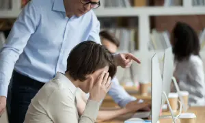 Przełożony wskazuje palcem na pracownicę, która zakrywa twarz dłońmi. Widać niewłaściwe zachowanie przełożonego wobec pracownika.