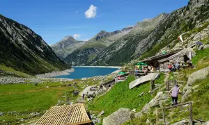 Ein idyllisches Bergpanorama mit einem türkisfarbenen See, umgeben von steilen Hängen. Hütten und Wanderer beleben das Bild, das an ein kleines Tibet im Zillertal erinnert.