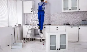 Un homme en salopette bleue est sur un escabeau, prêt à fixer un meuble au mur. La cuisine est en cours d'installation.