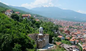 Eine malerische **Kosovo Landschaft** mit einer alten Kirche im Vordergrund, umgeben von üppigem Grün. Im Hintergrund erstreckt sich eine Stadt mit roten Dächern bis zu schneebedeckten Bergen.