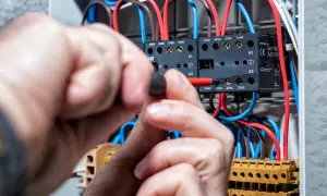 Ręce majsterkowicza przykręcają śrubę w skrzynce elektrycznej, gdzie widać przewody i styczniki. Kontrola napięcia prądu.