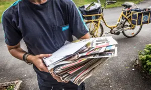Postnord-utdelare med post i händerna, redo för postutdelning varannan dag. En gul postcykel syns i bakgrunden.