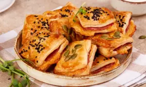 Pizzerinki z ciasta francuskiego, posypane czarnuszką i pestkami dyni, ułożone na talerzu. Idealne na przekąskę.