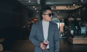 Mann im grauen Anzug mit weißem Rollkragenpullover und Sonnenbrille, hält Getränk. Das ist smart casual.
