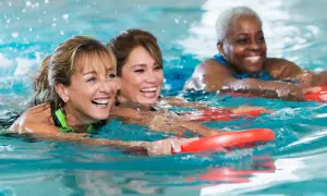 Three women joyfully participate in an adult learn to swim class, holding red kickboards as they move through the water.