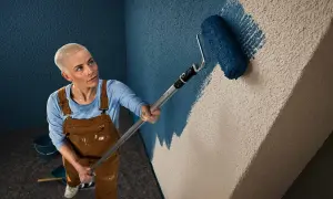 Frau mit Kurzhaarschnitt streicht mit einer Farbrolle eine Wand mit Rauputz. Sie trägt Latzhose und blaue Langarmshirt.