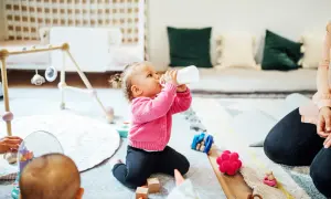 Mała dziewczynka pije z butelki, zastanawiając się, jak długo może stać mleko matki w podgrzewaczu. Wokół zabawki i inne dzieci.