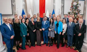 Grupa osób w eleganckich strojach pozuje do zdjęcia na tle flag i choinki.