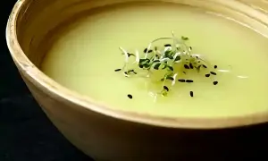 Un bol en bois contient un velouté d'asperges blanches, garni de pousses vertes et de graines noires.