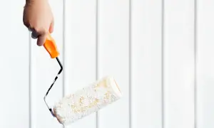 En hand håller en färgroller redo för målning utomhus mot en vit panelvägg.
