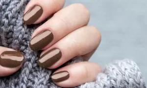 Dłonie z perfekcyjnym manicurem kombinowanym w odcieniu czekolady, otulone miękkim, szarym swetrem.