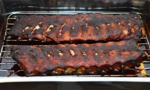 Zwei Rippchen-Stränge, dunkelbraun und saftig, ruhen auf einem Rost in einer Bratpfanne. Sie sind perfekt gegart, wie lange Spareribs im Ofen sein sollten.