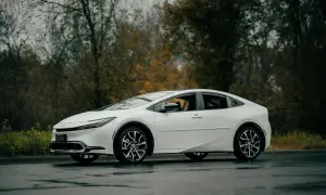 Biała Toyota Prius zaparkowana na mokrej drodze, z drzewami w tle.