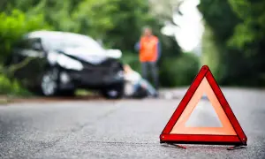 Triangle de signalisation sur la route, près d'une voiture accidentée où une personne gît au sol. Un homme en gilet orange se tient debout. Scène évoquant un homicide involontaire.