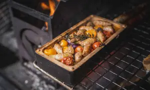 Salsiccia mit Gemüse in einer Auflaufform auf dem Grill, Flammen im Hintergrund.