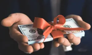 Dłonie trzymają plik banknotów 100-dolarowych przewiązany czerwoną wstążką. To pieniądze zamiast prezentów na urodziny.