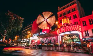 Nocny Paryż rozświetlony neonami Moulin Rouge. Wirujący wiatrak i czerwone światła tworzą magiczną atmosferę.