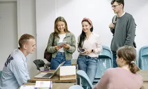 Grupa młodych ludzi dyskutuje o tym, ile trwa liceum dla dorosłych, przeglądając notatki i książki przy drewnianym stole.