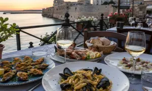Un tavolo con vista sul mare, pronto per gustare squisiti piatti tipici tarantini: pasta con cozze, soutè di cozze e un misto di crostacei.